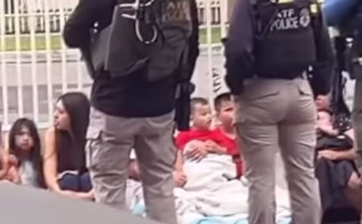 two officers stand in front of a row of women and kids sitting on the side of the road